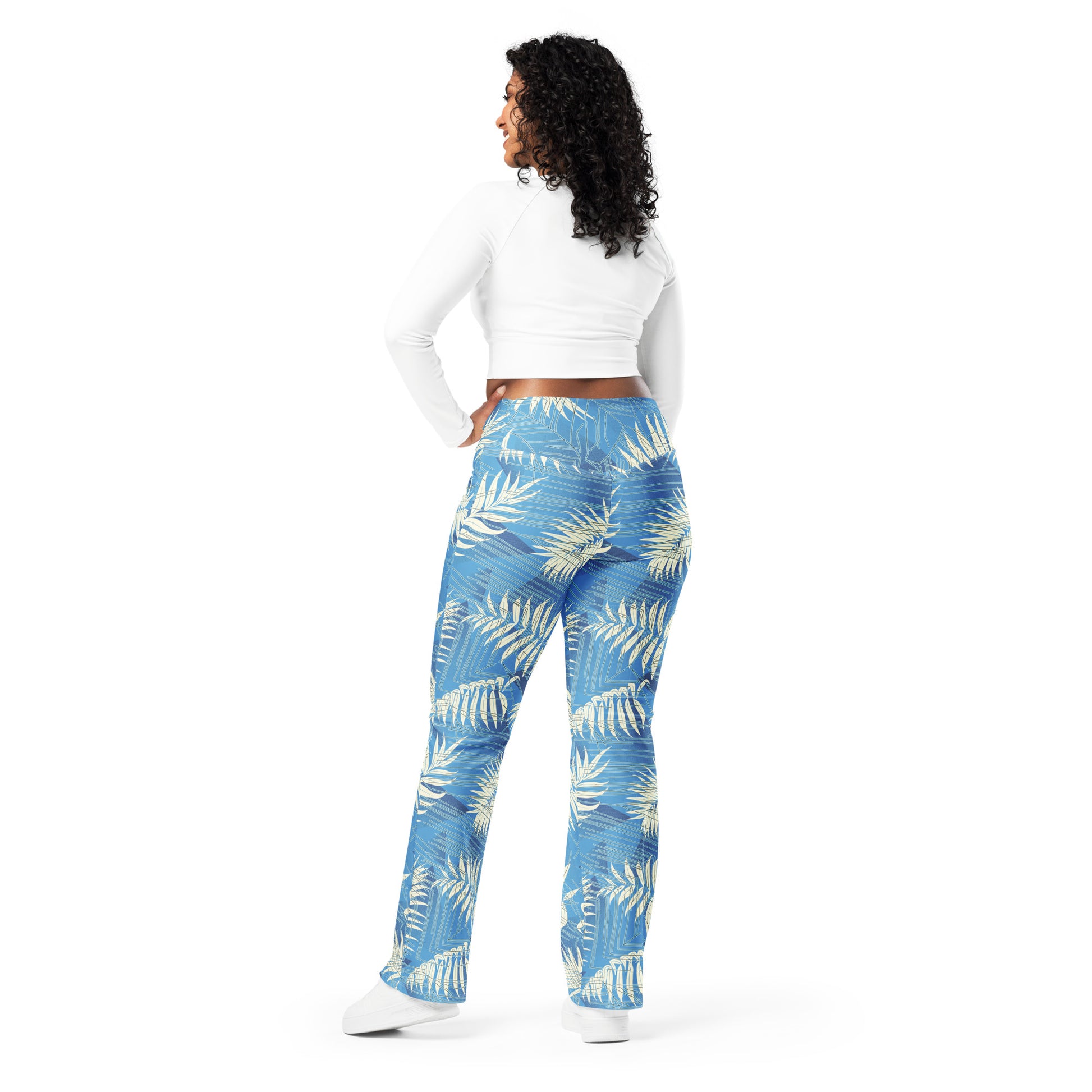 A woman shown from the back wearing blue pants with a white palm leaf pattern, a white long-sleeve crop top, and white sneakers, set against a plain white background. Bold Contemporary Tropical Palm Leaves Flare Leggings
