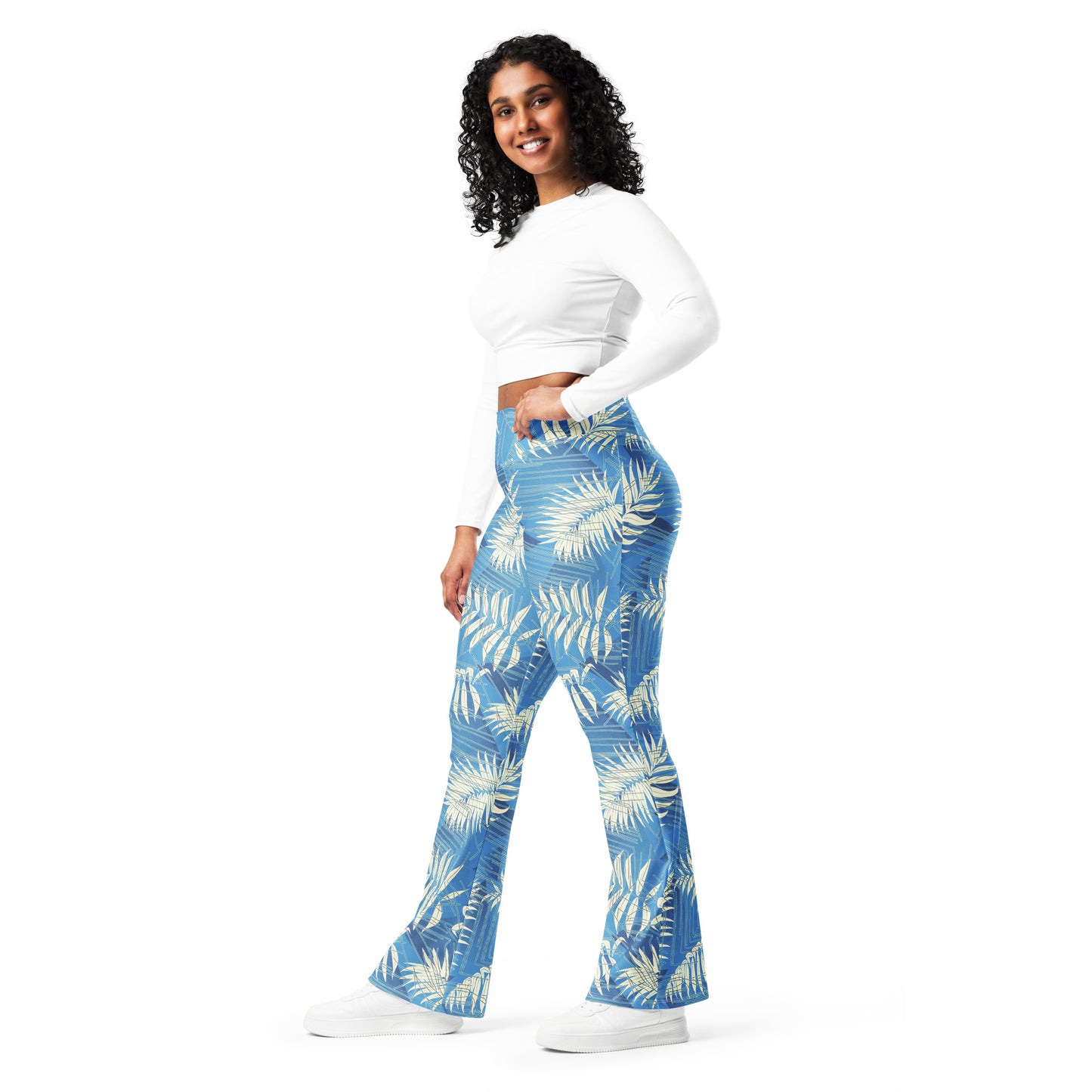 A woman modeling a pair of high-waisted, wide-leg pants with a blue and cream palm leaf pattern, paired with a white long-sleeve crop top and white sneakers, set against a plain white background. Bold Contemporary Tropical Palm Leaves Flare Leggings Activewear
