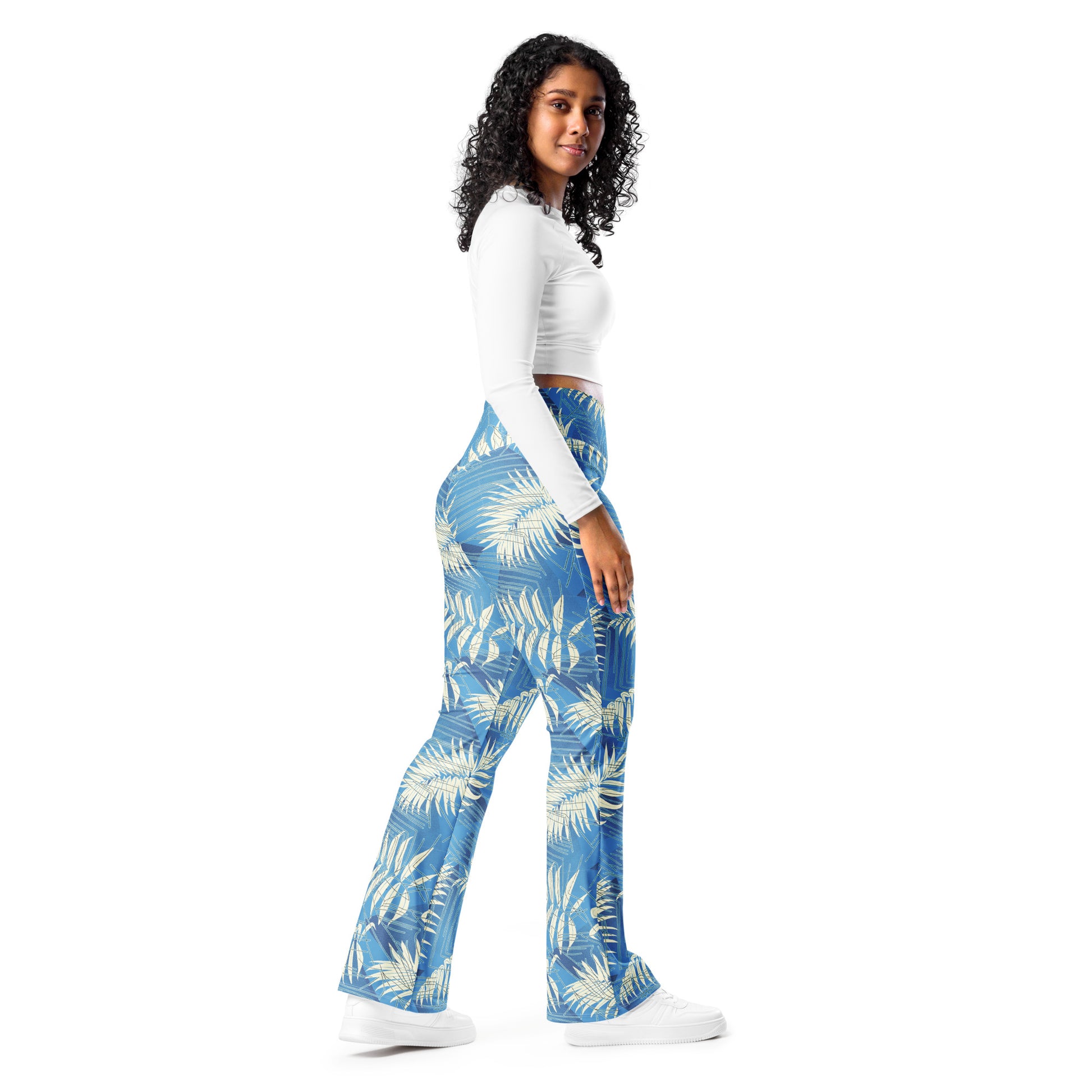 A woman modeling high-waisted, wide-leg pants with a blue and cream palm leaf pattern, paired with a white long-sleeve crop top and white sneakers, set against a plain white background. Bold Contemporary Tropical Palm Leaves Flare Leggings for Activewear