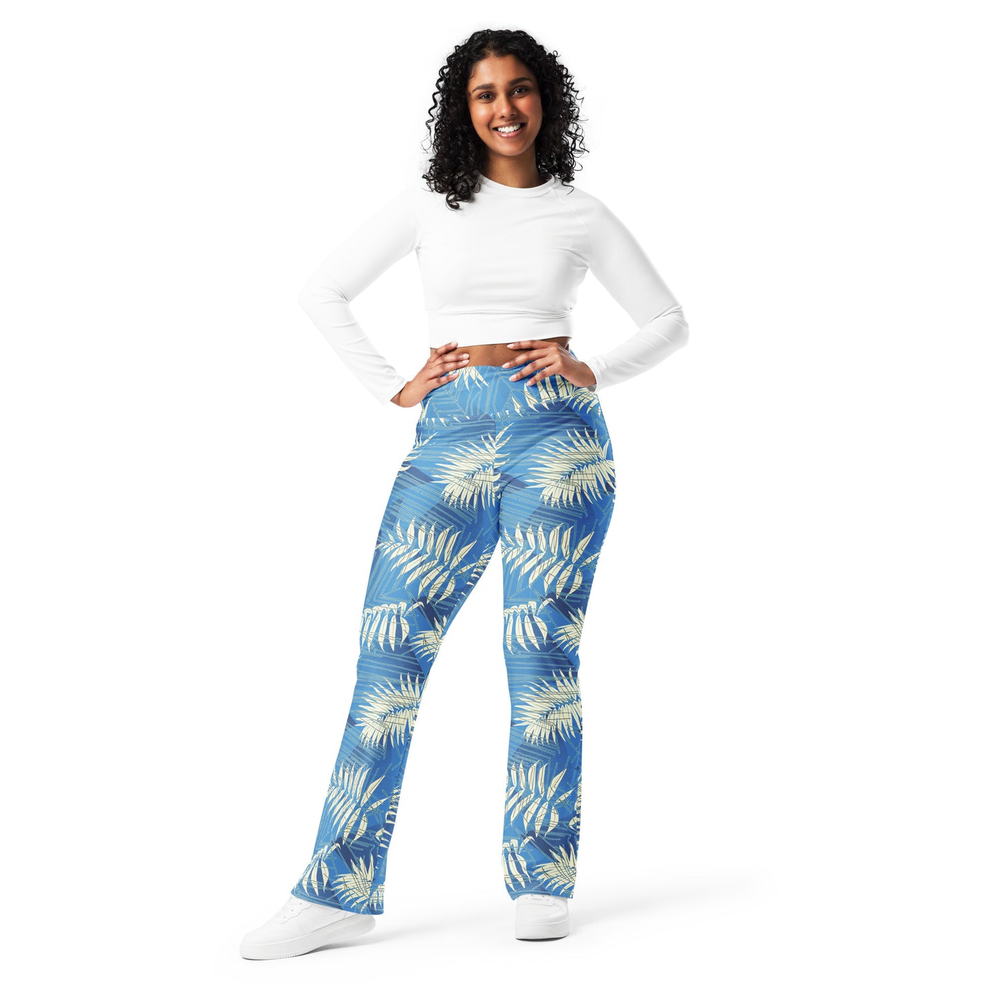 A woman modeling high-waisted blue pants with a cream palm leaf pattern, paired with a white long-sleeve crop top and white sneakers, against a solid white background. Contemporary Tropical Palm Leaves Flare Leggings