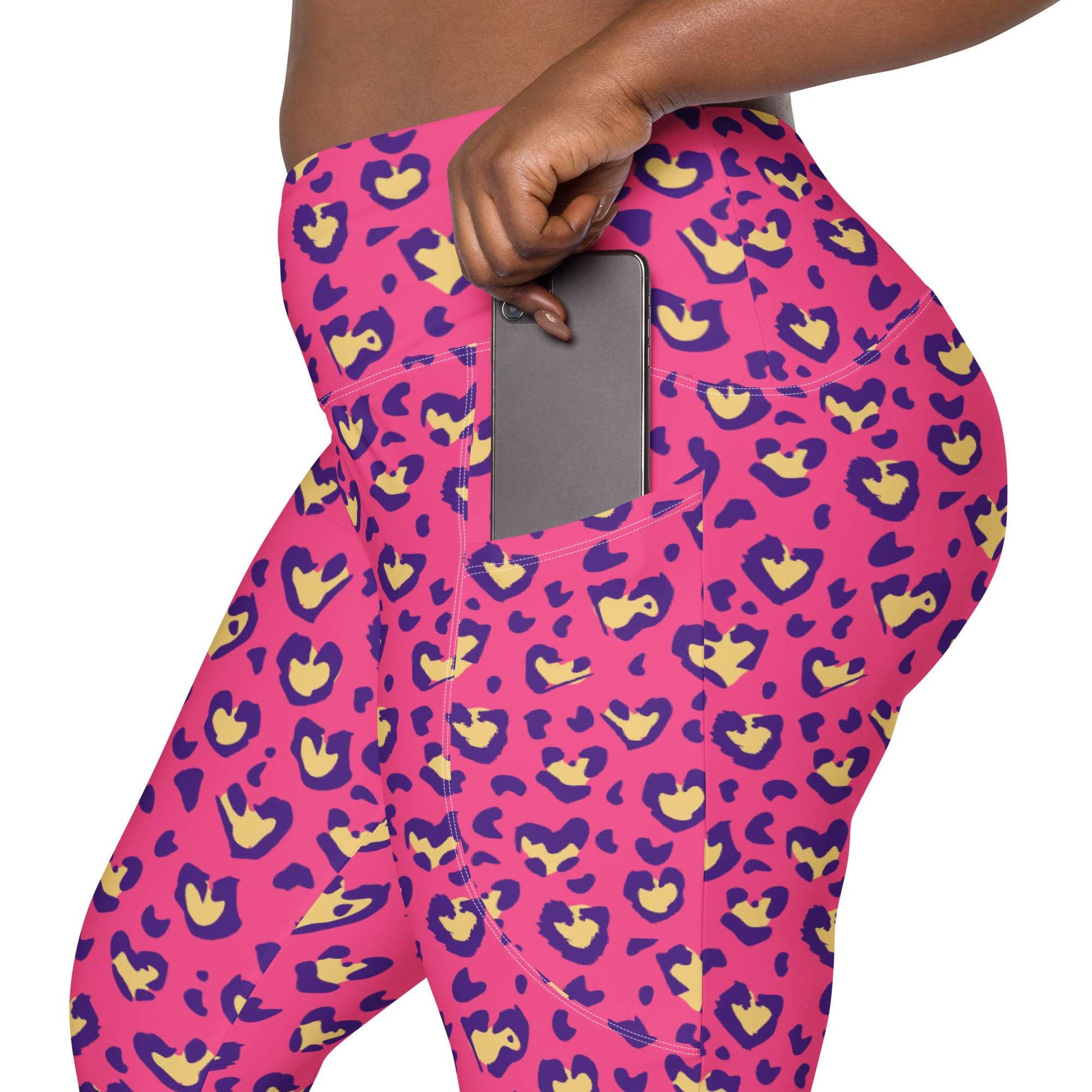 Close-up of a person wearing bright pink leggings with a purple and yellow abstract pattern, shown from the waist to upper thigh. A hand is pulling up the side of the waistband, and a gray smartphone is tucked into the side pocket. Heart Pink Leopard Pattern Leggings with Pockets Activewear Shop Heart Pink Leopard Pattern Leggings with Pockets Activewear, designed for bold workouts, everyday comfort, and convenient storage. Upgrade your workout wardrobe with Heart Pink Leopard Pattern Leggings with Pockets