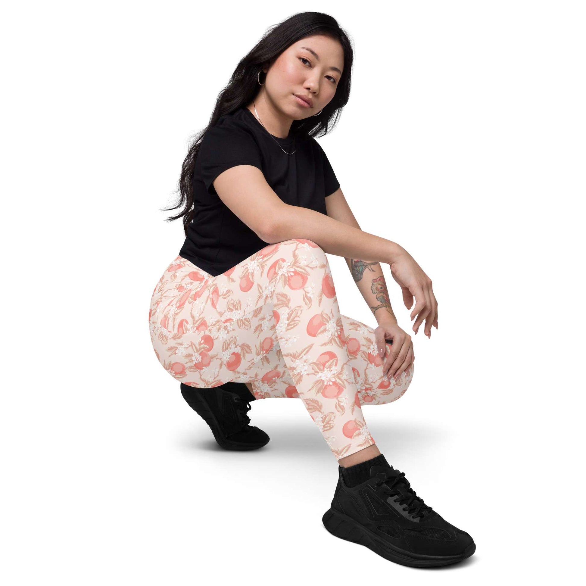 A woman crouching in a studio setting, wearing black athletic shoes, a black short-sleeve t-shirt, and patterned leggings with a peach and pink floral design on a cream background. Artistic Ditsy Floral Print Leggings with Pockets Activewear Artistic Stylized Ditsy Floral Pattern with Blue and Orange Botanical Motifs Leggings with Pockets Activewear Shop Artistic Ditsy Floral Print Leggings with Pockets, designed for bold outfits, flexible movement, and convenient everyday storage.